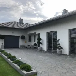 Nowoczesny dom jednorodzinny z szarą bramą garażową, żaluzjami zewnętrznymi i brukowanym podjazdem, otoczony zielenią i ozdobnymi drzewkami.