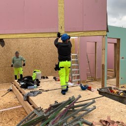 Inster Group Sp. z o.o. - Dwóch pracowników w odblaskowych ubraniach montuje ściany z płyt OSB i różowej wełny mineralnej wewnątrz powstającego budynku.