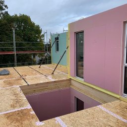 Inster Group Sp. z o.o. - Budowa domu: widok z góry na drewnianą konstrukcję stropu z wyciętym otworem, różowe panele izolacyjne na ścianach, okna wstawione, w tle rusztowanie i drzewa.