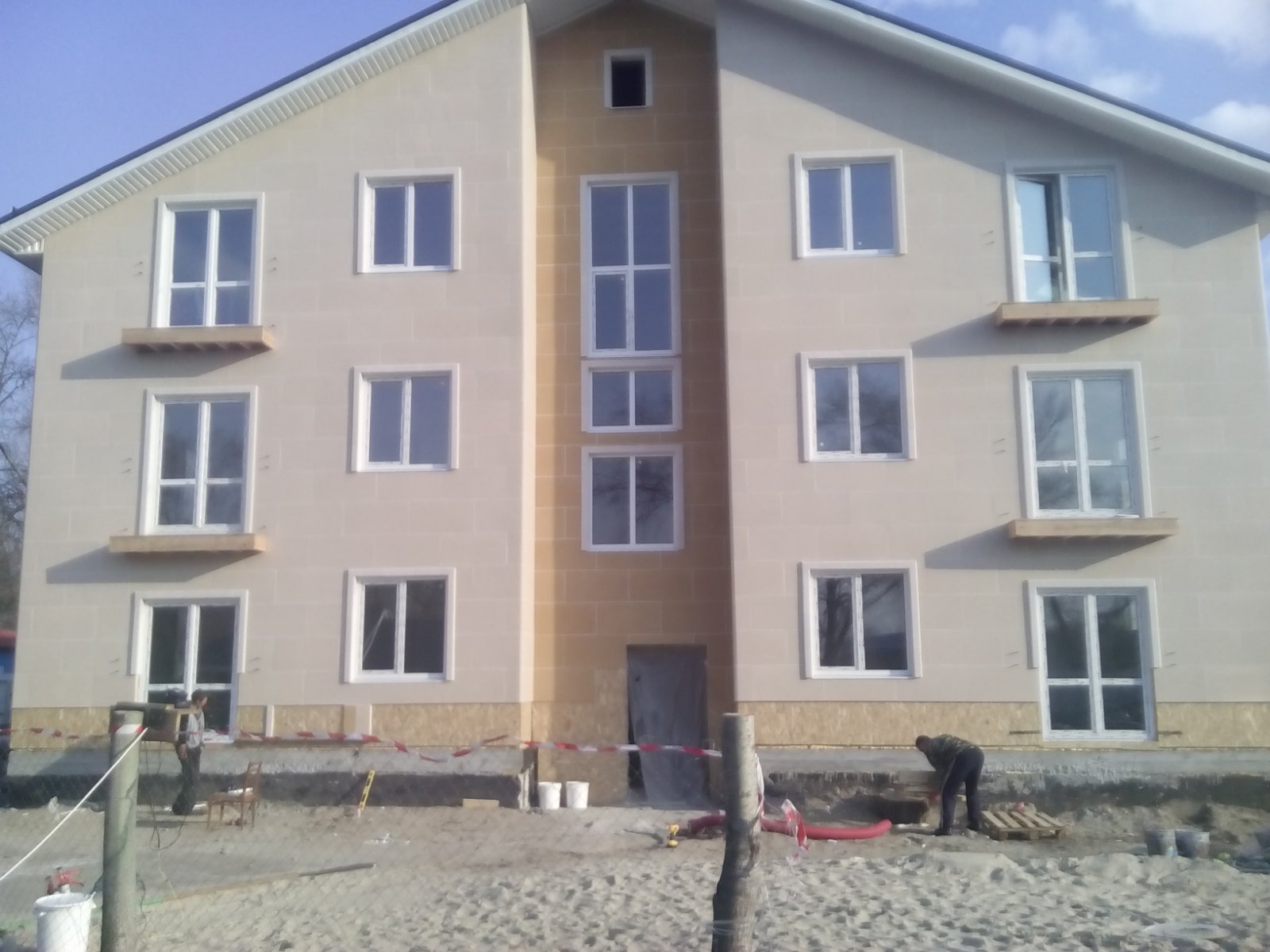 Dwupiętrowy dom w trakcie budowy z widocznymi pracownikami na zewnątrz, jasna elewacja z białymi ramami okiennymi i drewnianymi elementami pod oknami, teren budowy otoczony taśmą.