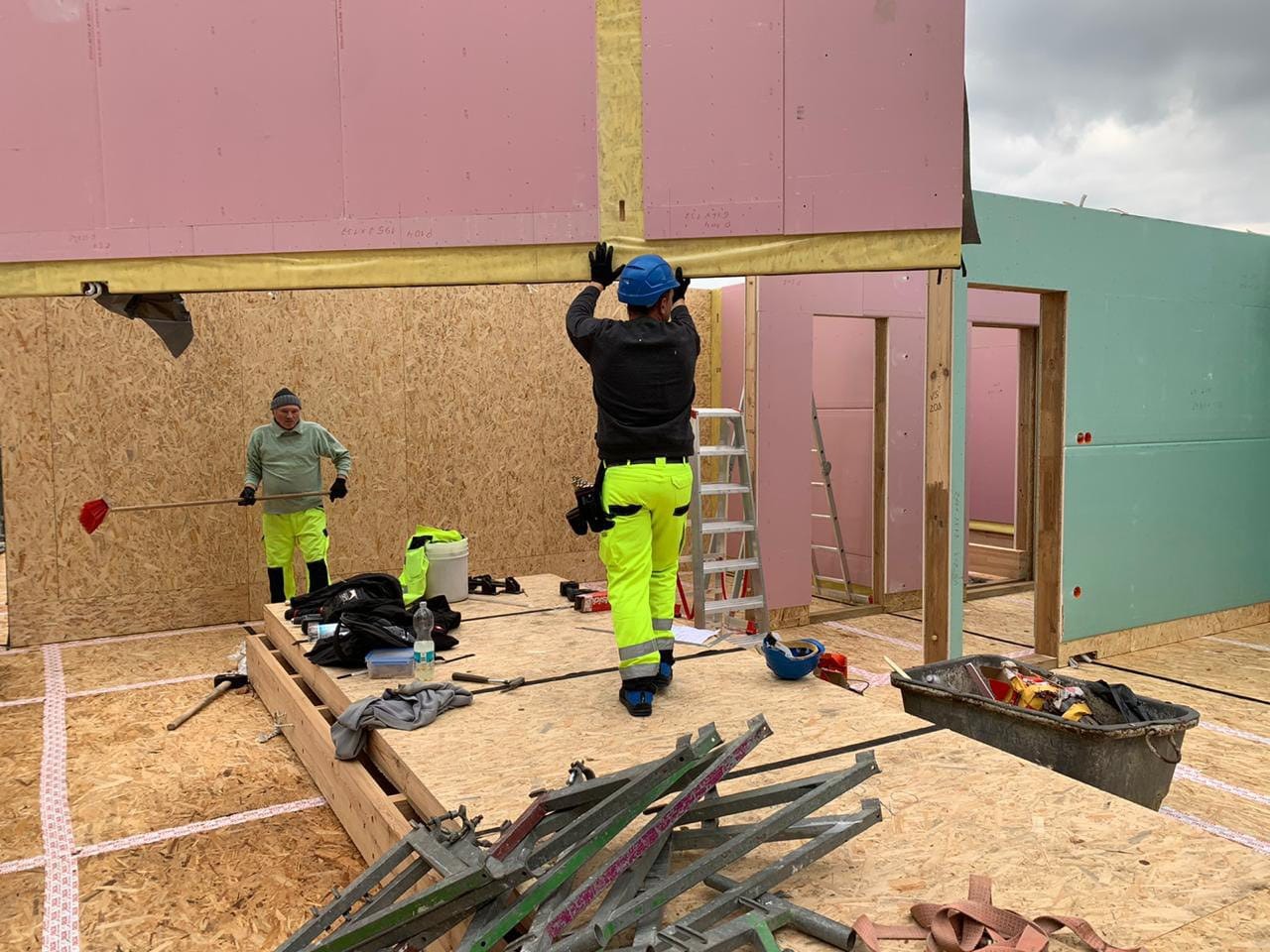 Dwóch pracowników w odblaskowych ubraniach montuje ściany z płyt OSB i różowej wełny mineralnej wewnątrz powstającego budynku.