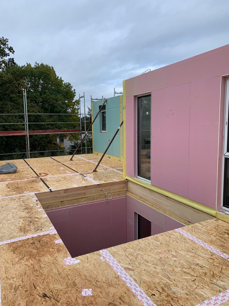 Budowa domu: widok z góry na drewnianą konstrukcję stropu z wyciętym otworem, różowe panele izolacyjne na ścianach, okna wstawione, w tle rusztowanie i drzewa.