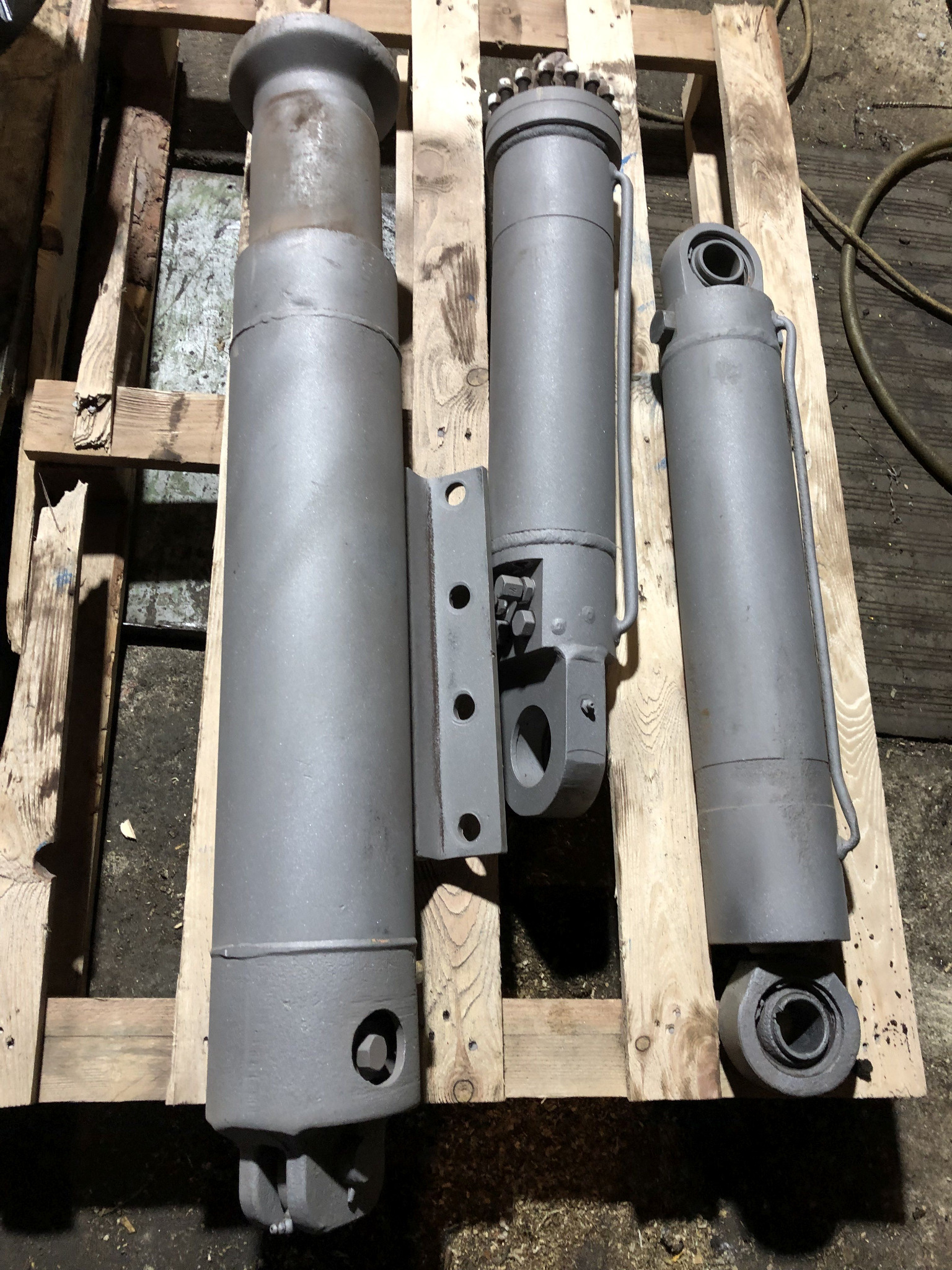 Trzy cylindry hydrauliczne po piaskowaniu i malowaniu proszkowym, leżące na drewnianej palecie, widoczne detale mocowań i otworów.