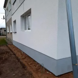 Świeżo otynkowana elewacja domu jednorodzinnego z widocznym fragmentem starej elewacji, przygotowana do dalszych prac wykończeniowych w gruncie.