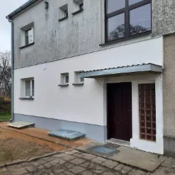 Dwukondygnacyjny budynek z częściowo odnowioną elewacją, gdzie górna część pozostaje w szarym tynku, a dolna została pomalowana na biało, z widocznym zadaszeniem nad wejściem i kostką brukową przed...