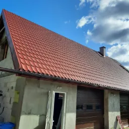 Dom w stanie surowym z nowym, czerwonym dachem z blachodachówki, widoczne rynny, okno dachowe i komin, obok garaż z brązową bramą segmentową, na ziemi resztki blachodachówki i niebieski kosz...