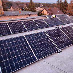 SUN NRG PRO sp.z.o.o Fotowoltaika - Instalacja paneli słonecznych na płaskim dachu budynku, widoczny pracownik w żółtej kamizelce odblaskowej, w tle zabudowa jednorodzinna i drzewa.