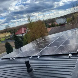 Panele słoneczne zamontowane na metalowym dachu z widocznym kominkiem wentylacyjnym, w tle zabudowa jednorodzinna i pochmurne niebo.