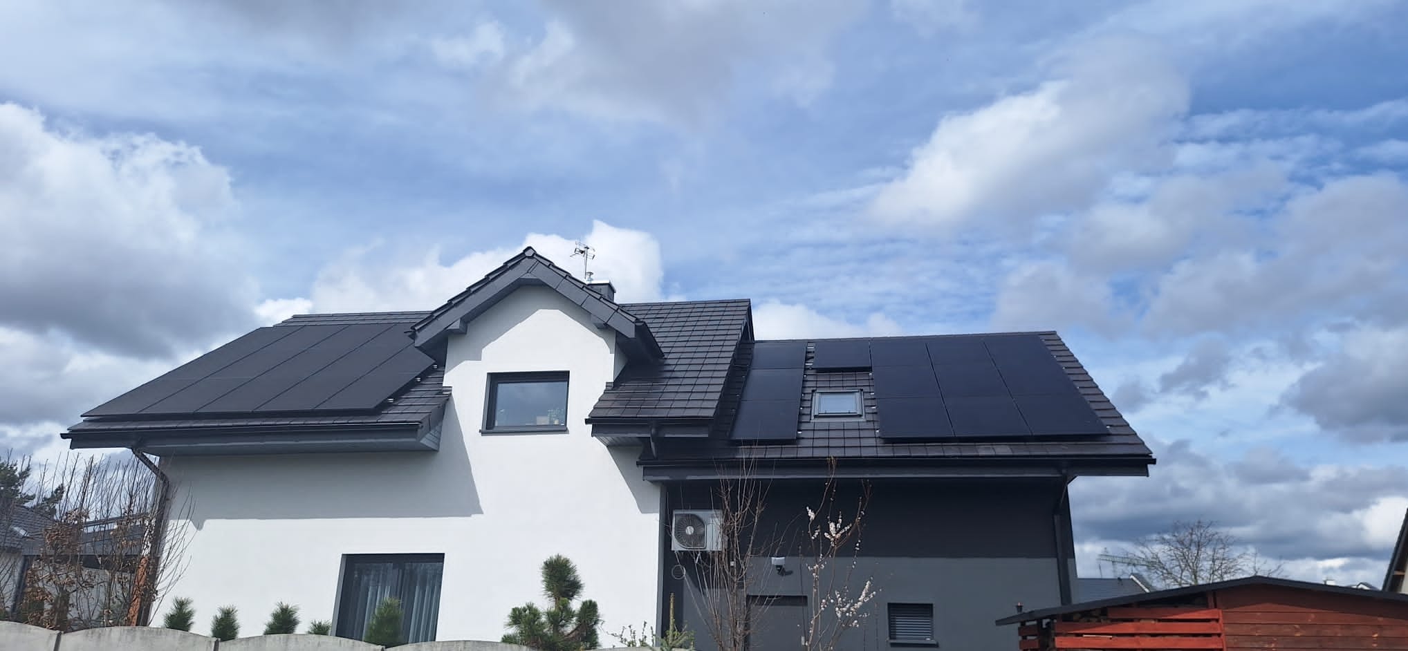 Dwukondygnacyjny dom z szarą dachówką i panelami fotowoltaicznymi na dachu, widok z dołu na tle pochmurnego nieba.