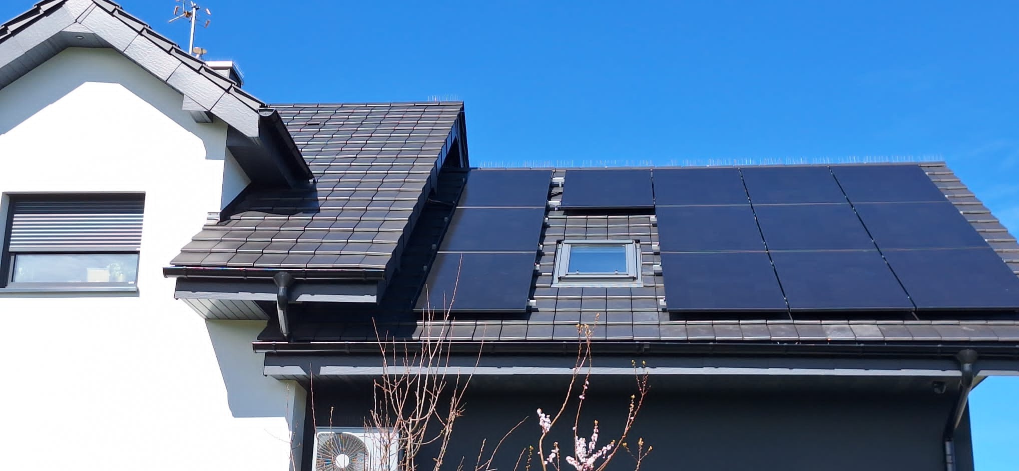 Ciemne panele słoneczne zamontowane na szarej dachówce nowoczesnego domu jednorodzinnego z oknem dachowym, widoczne kolce przeciw ptakom.