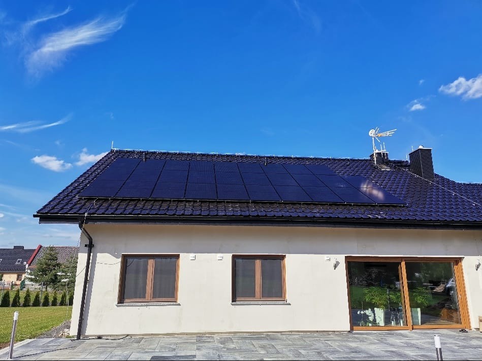 Panele fotowoltaiczne na dachu domu jednorodzinnego z jasną elewacją, widoczne okna z brązowymi ramami i przesuwne drzwi tarasowe, błękitne niebo z lekkimi obłokami w tle.
