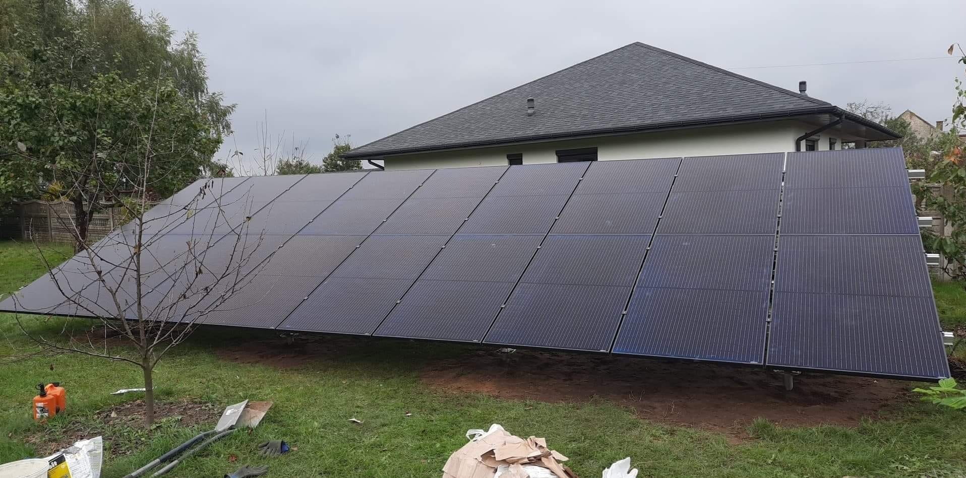 Panele słoneczne zamontowane na ziemi przed domem z szarym dachem, widoczne narzędzia i materiały budowlane, drzewo bez liści po lewej stronie.