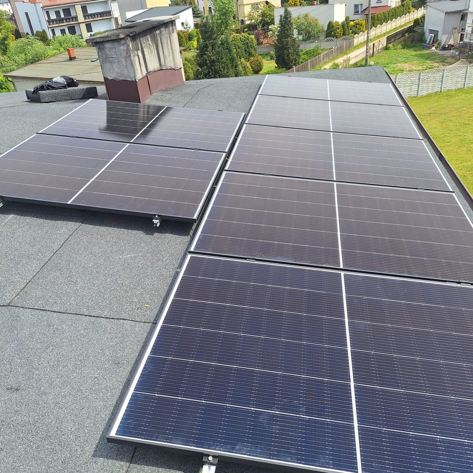 Panele słoneczne zamontowane na płaskim dachu pokrytym papą, widok z góry, fragment komina w tle.