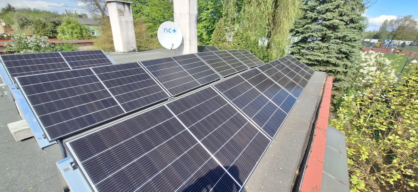 Panele słoneczne na dachu budynku z widoczną anteną satelitarną i zielenią w tle.