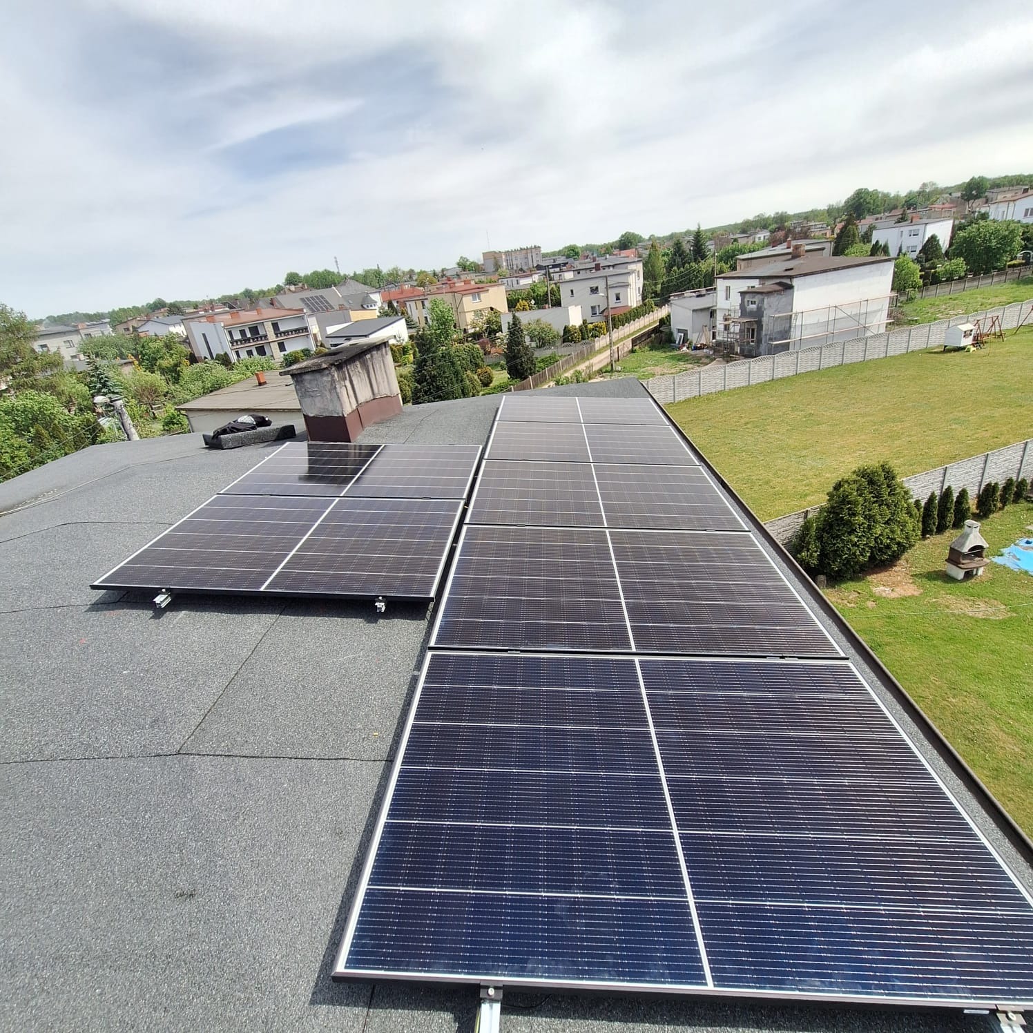 Panele słoneczne na płaskim dachu pokrytym papą, widok z góry na tle zabudowy jednorodzinnej i zielonego trawnika.