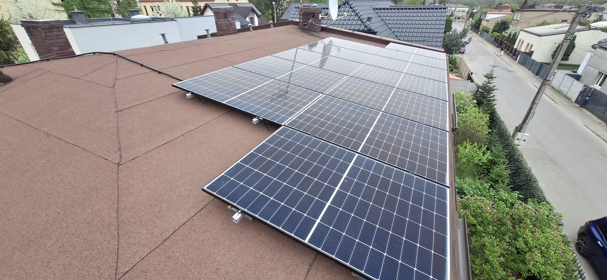 Instalacja fotowoltaiczna na dachu pokrytym papą, widok z góry na panele odbijające niebo, z widoczną ulicą i zabudową w tle.