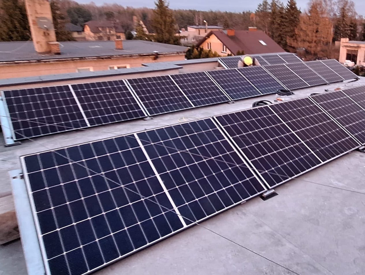 Instalacja paneli słonecznych na płaskim dachu budynku, widoczny pracownik w żółtej kamizelce odblaskowej, w tle zabudowa jednorodzinna i drzewa.