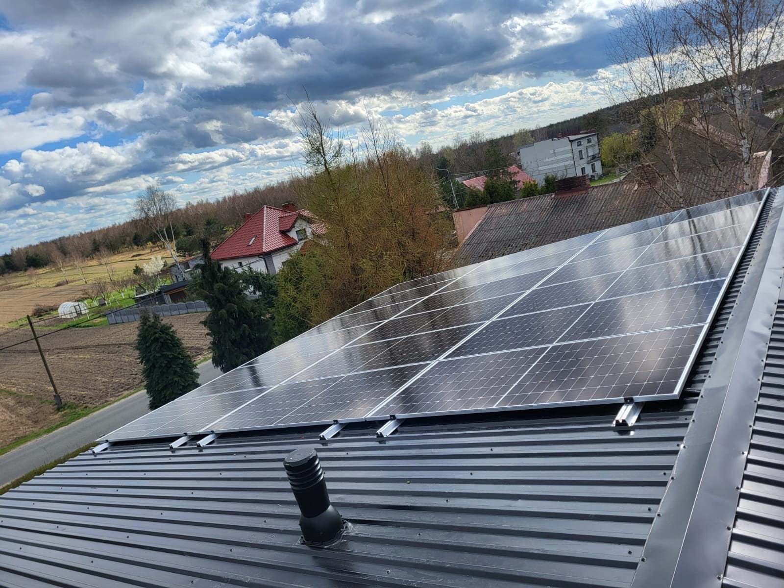 Panele słoneczne zamontowane na metalowym dachu z widocznym kominkiem wentylacyjnym, w tle zabudowa jednorodzinna i pochmurne niebo.