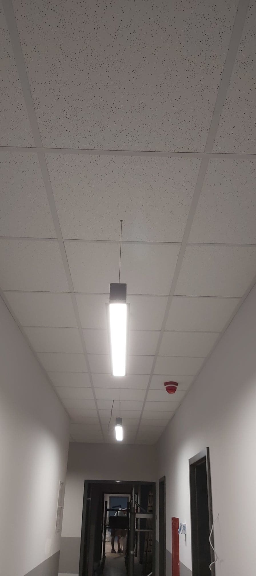 Sufit podwieszany z oświetleniem rastrowym w korytarzu biurowym w Toruniu, widoczny czujnik dymu.