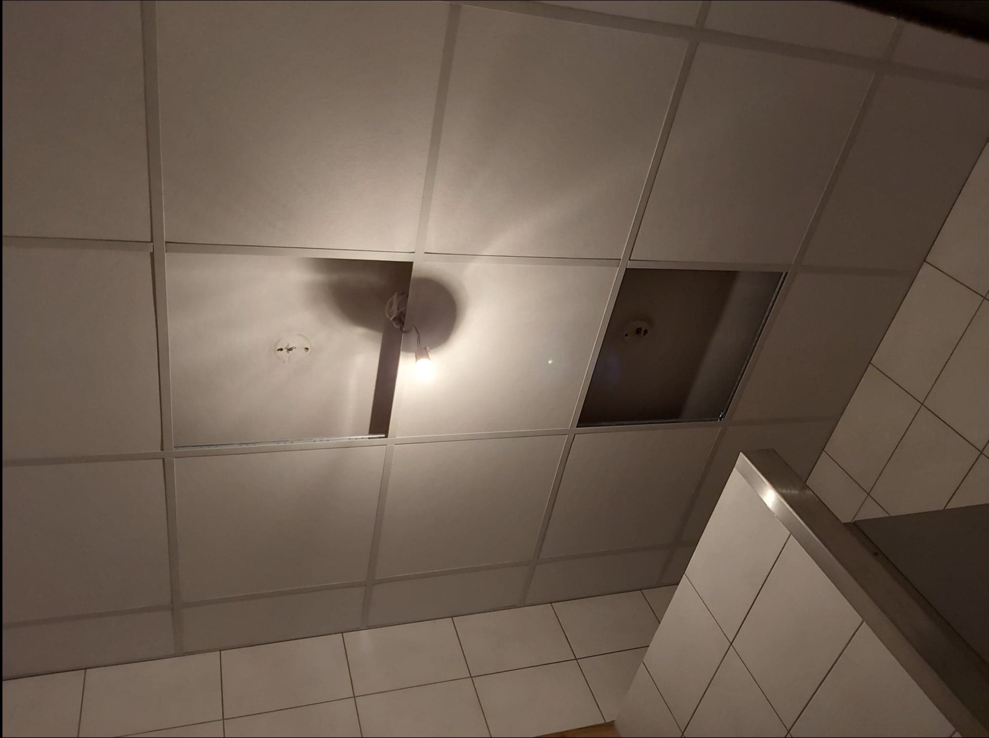 Uszkodzony sufit podwieszany z odsłoniętą instalacją i brakiem kilku paneli, widoczne płytki ceramiczne na ścianie.