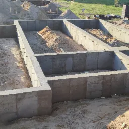 Wylane betonowe fundamenty domu jednorodzinnego z wypełnieniem piaskiem wewnątrz, widoczne zbrojenia, w tle pracownicy budowlani i ułożone bloczki