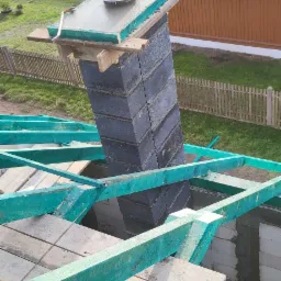 Wznoszenie komina z szarych bloczków na budowie domu, widoczne zielone krokwie i fragment dachu z membraną, w tle budynek i ogrodzenie.