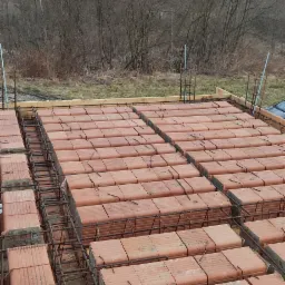 Wieniec stropowy z cegieł ceramicznych i zbrojenia stalowego, widok z góry, w tle drzewa bez liści i pochmurne niebo.