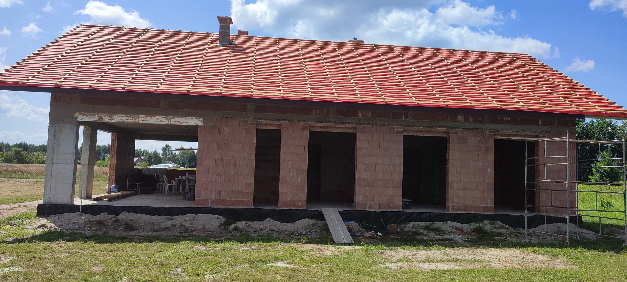 Surowy stan domu z czerwonej cegły z przygotowaną konstrukcją pod dachówkę, widoczne rusztowanie i teren budowy.