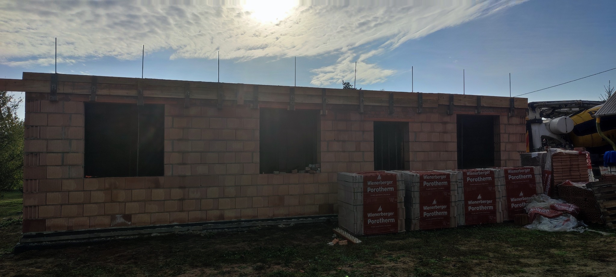 Wznoszenie ścian zewnętrznych domu jednorodzinnego z czerwonej cegły Porotherm, widoczne otwory okienne i drzwiowe, drewniane szalunki stropowe, w tle mikser do betonu.