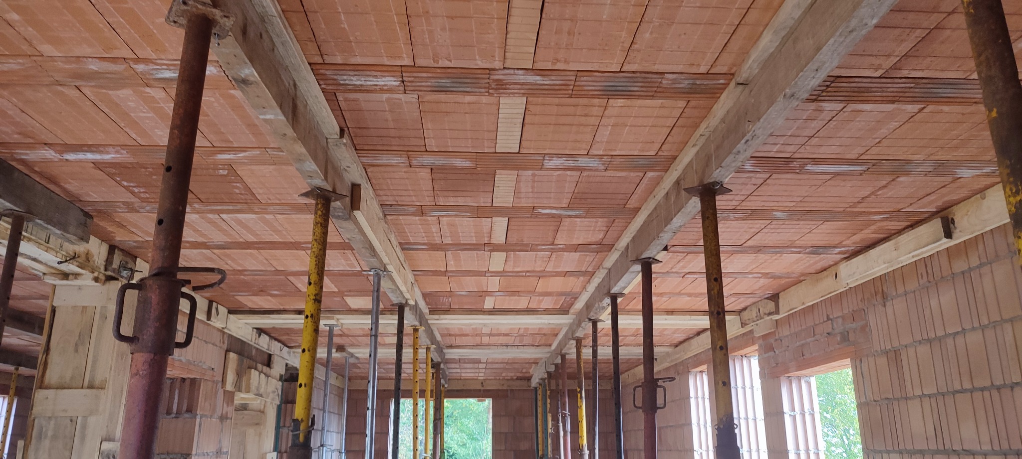 Wnętrze domu w trakcie budowy: widok na strop z pustaków ceramicznych podpartych stemplami budowlanymi, widoczne drewniane belki i murowane ściany.