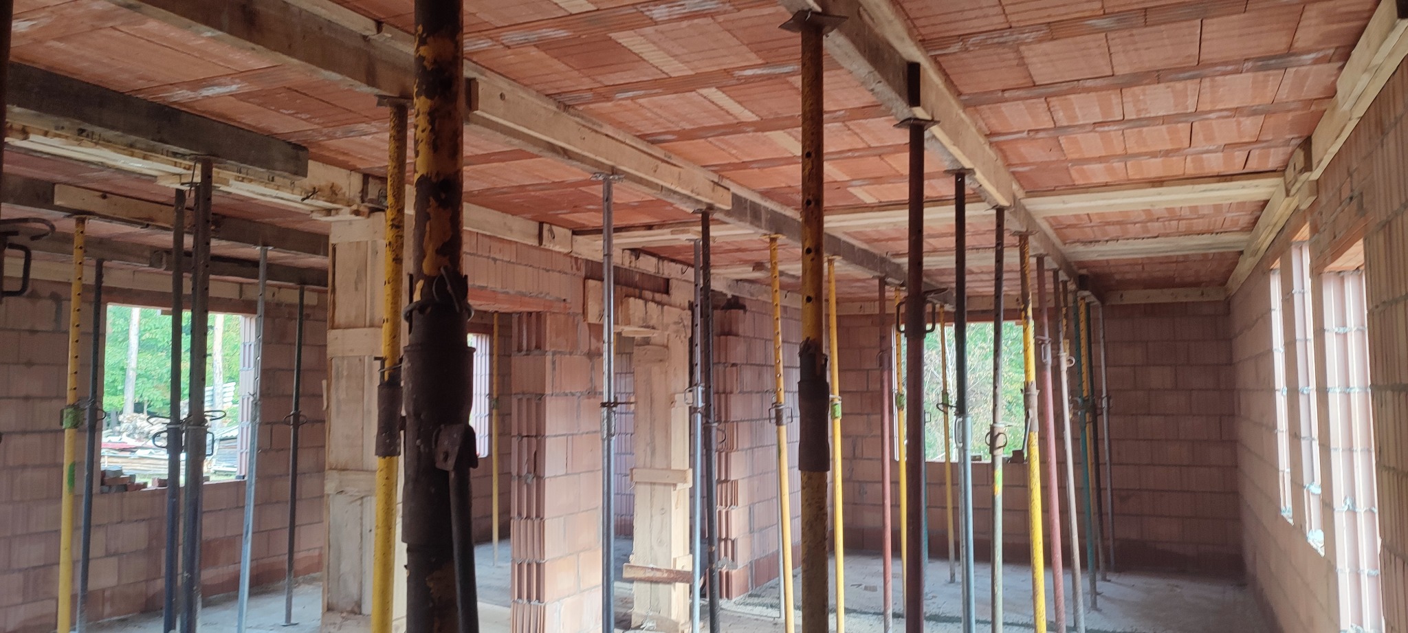 Wnętrze w trakcie budowy: ściany z czerwonej cegły, drewniane belki stropowe podparte metalowymi stemplami, widok na zewnątrz przez otwory okienne.