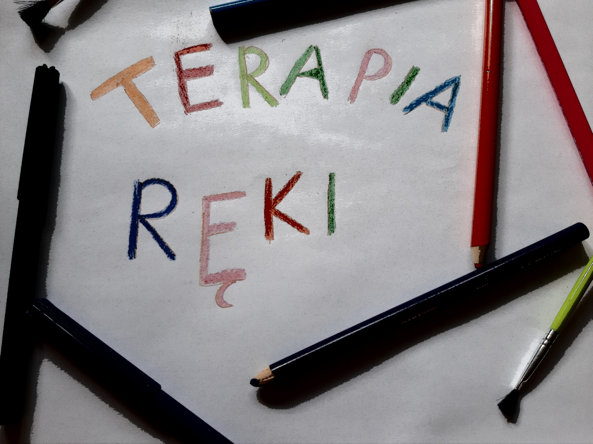 Napis 'TERAPIA RĘKI' wykonany kredkami w różnych kolorach, otoczony kredkami i pędzlami na białym tle.