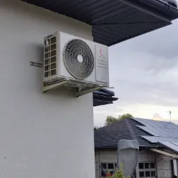 Biała jednostka zewnętrzna klimatyzacji marki Rotenso zamontowana na ścianie domu z szarą elewacją i ciemnym dachem, w tle widoczne panele słoneczne.