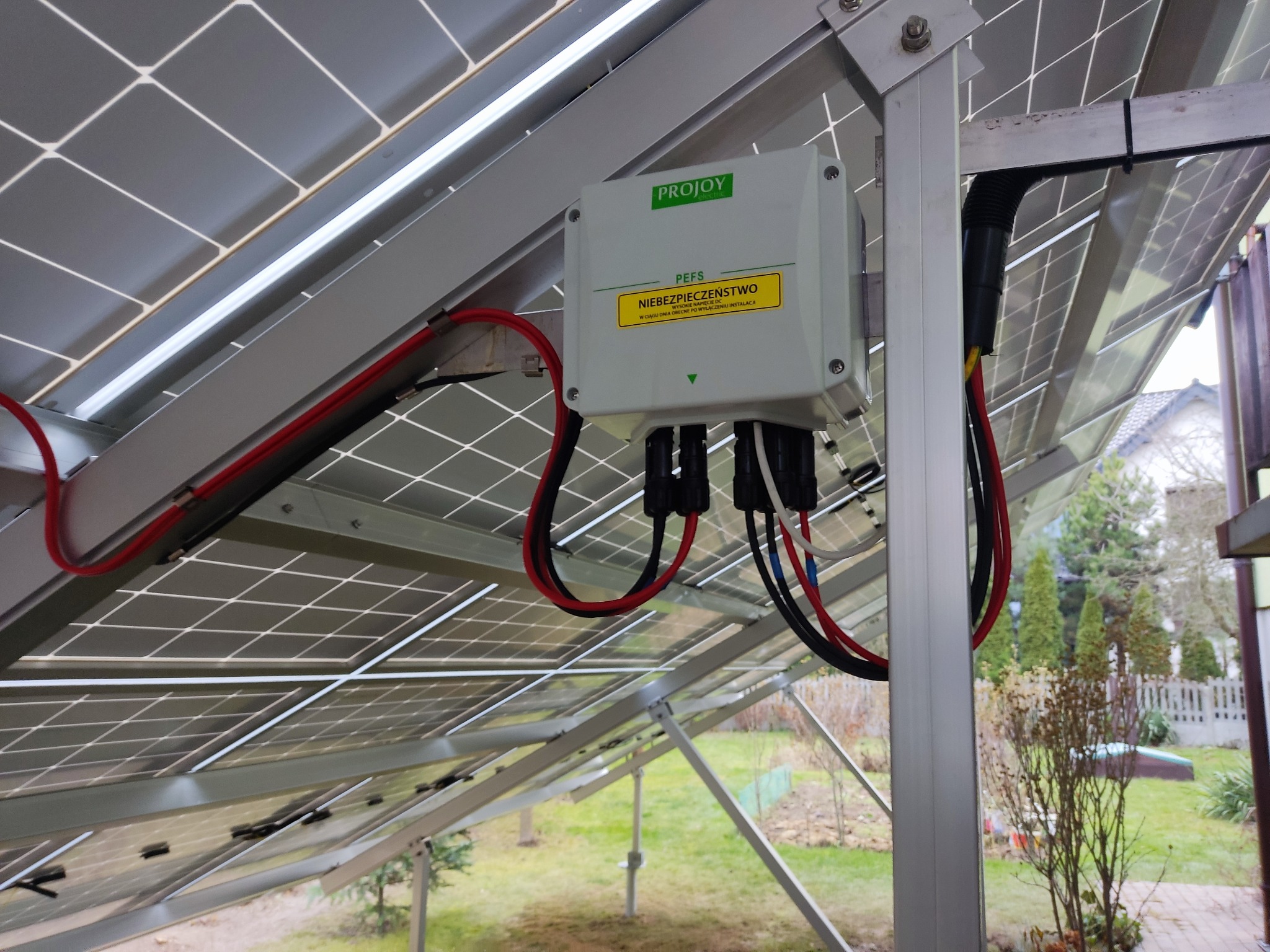 Szara skrzynka PEFS z żółtą naklejką ostrzegawczą 'NIEBEZPIECZEŃSTWO' podłączona do paneli fotowoltaicznych na metalowej konstrukcji, widoczne przewody i złącza.