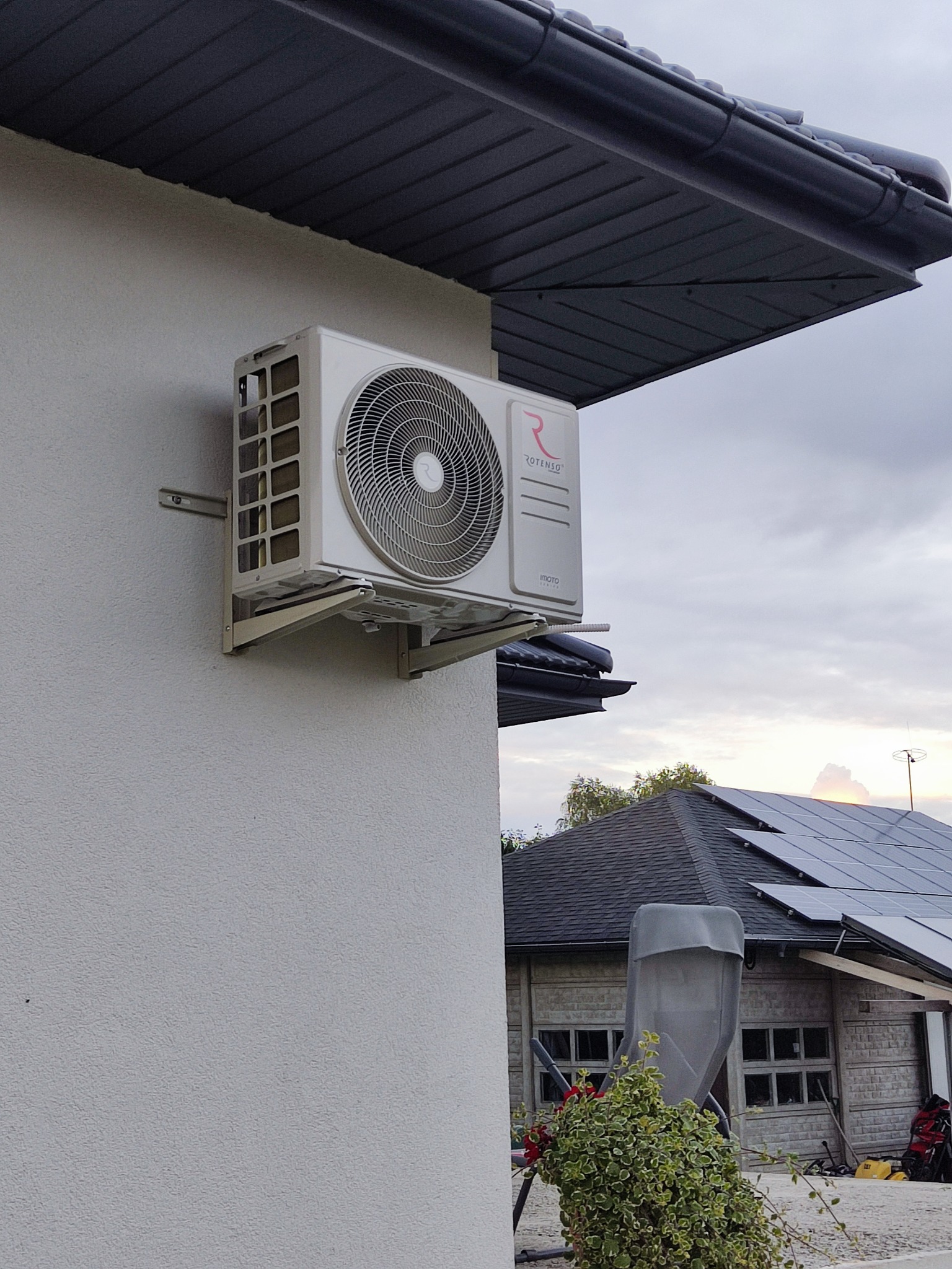 Biała jednostka zewnętrzna klimatyzacji marki Rotenso zamontowana na ścianie domu z szarą elewacją i ciemnym dachem, w tle widoczne panele słoneczne.
