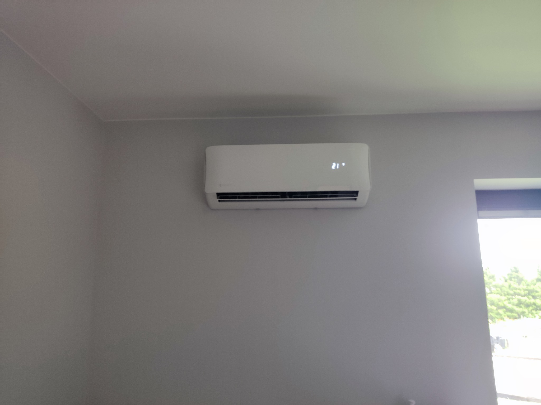 Biały klimatyzator ścienny z wyświetlaczem temperatury 21 stopni Celsjusza, zamontowany na szarej ścianie w jasnym pomieszczeniu z widokiem na zieleń za oknem.