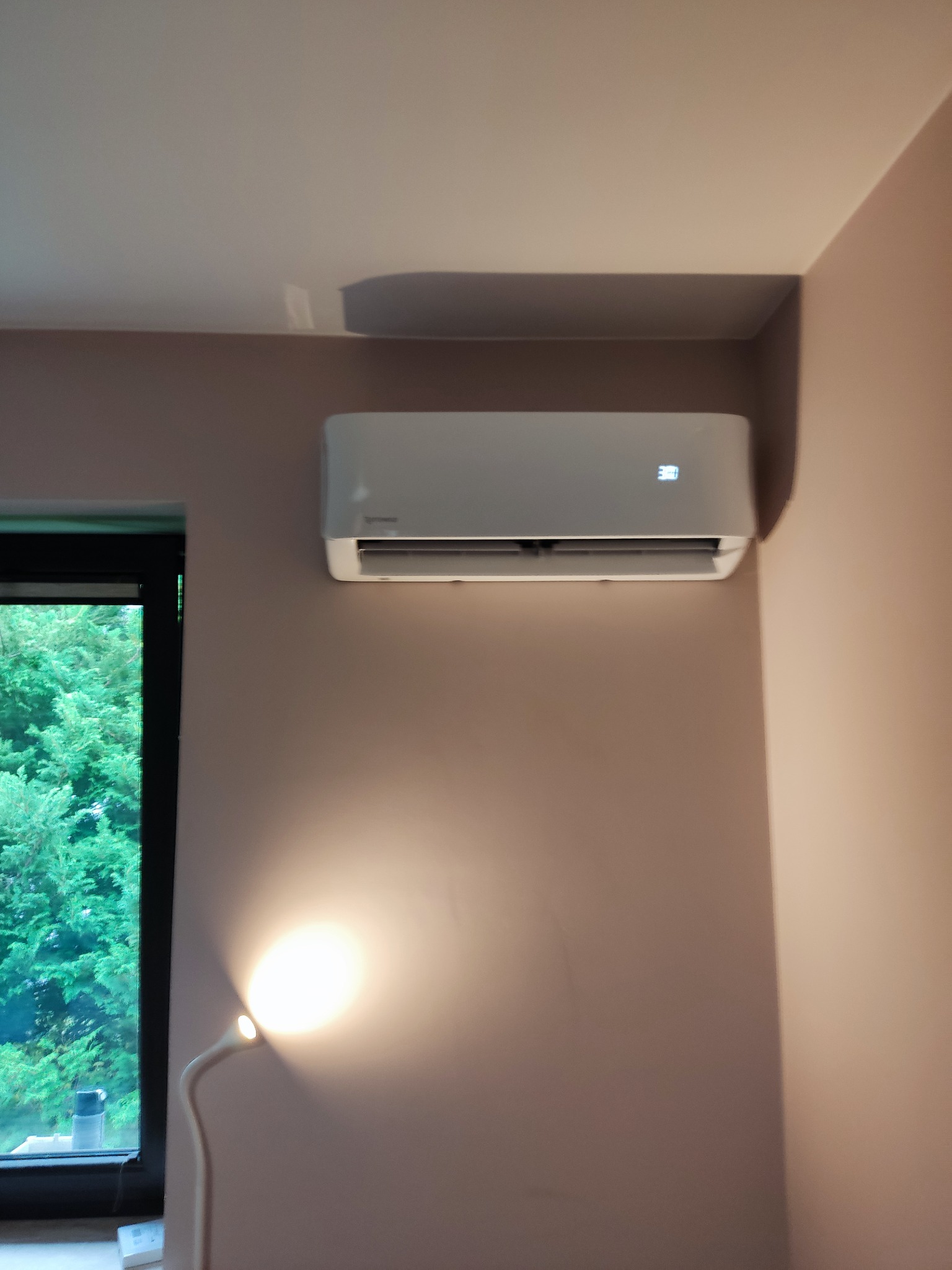 Biały klimatyzator ścienny marki Sprinco z wyświetlaczem temperatury 30 stopni, zamontowany w pokoju z oknem i lampką biurkową.