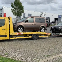 Żółta laweta marki Renault Master z brązowym Volkswagenem Caddy i czarnym Audi A6 Avant na przyczepie, na tle salonu samochodowego z logo Volkswagena.