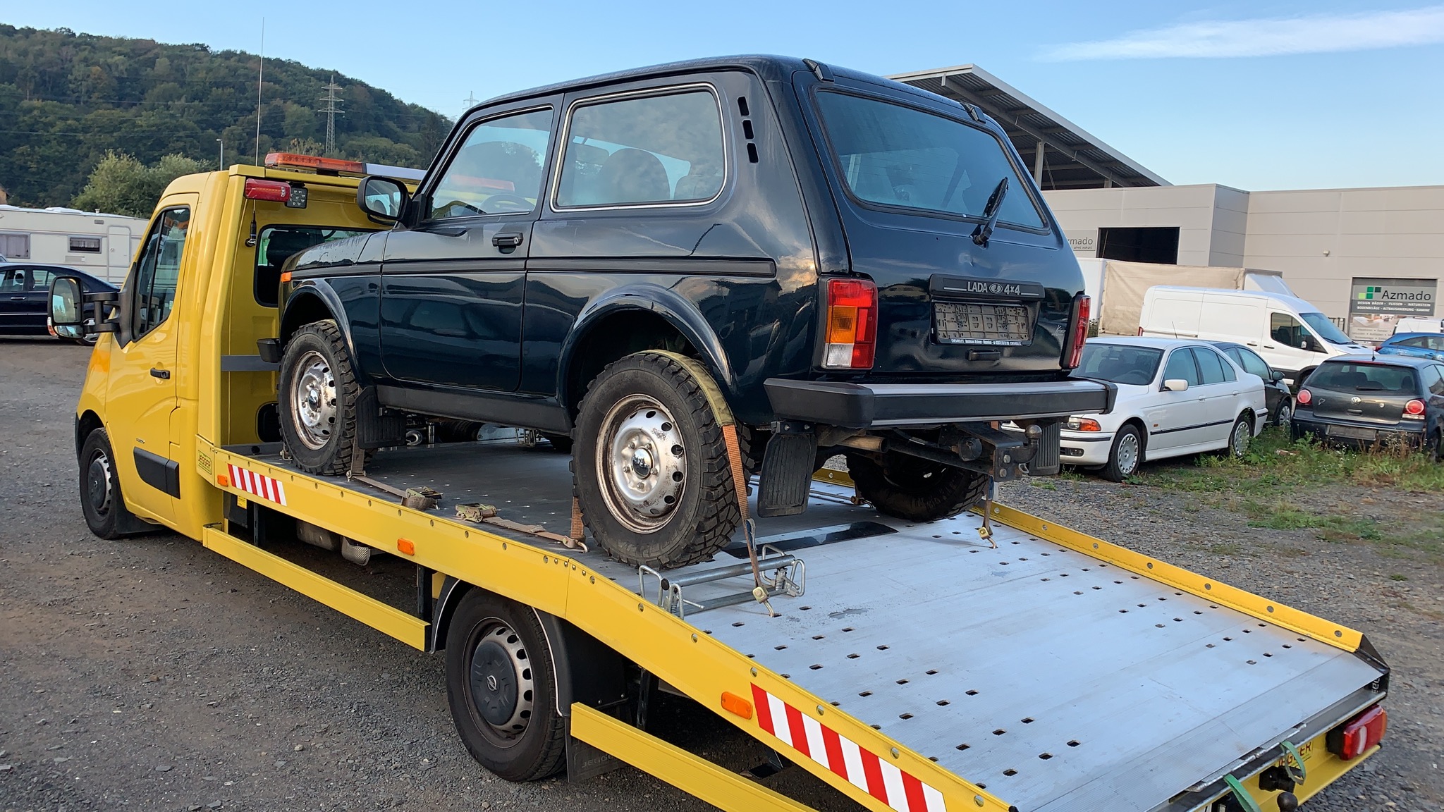 Czarna Lada Niva 4x4 zabezpieczona pasami na żółtej lawecie, w tle zabudowania i zieleń.