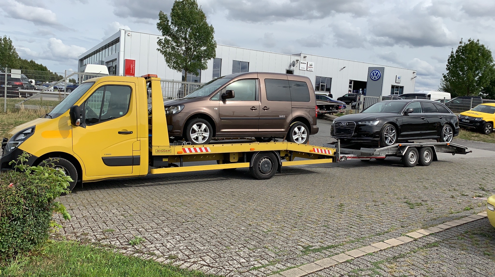 Żółta laweta marki Renault Master z brązowym Volkswagenem Caddy i czarnym Audi A6 Avant na przyczepie, na tle salonu samochodowego z logo Volkswagena.