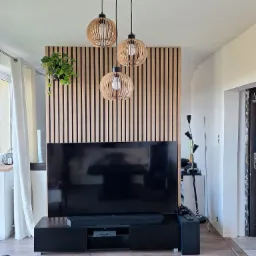 Nowoczesny salon z drewnianą ścianą z lameli za telewizorem, wiszącą rośliną, trzema lampami z drewnianymi abażurami i czarną szafką RTV.