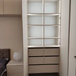 Wnętrze szafy z półkami i szufladami, oświetlenie LED wewnątrz, widok na szafkę nocną i łóżko w tle.