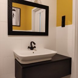 NeoMeb - Nowoczesna łazienka z białą umywalką nablatową, czarną szafką pod umywalką, lustrem w czarnej ramie z fakturą oraz żółtą ścianą w tle.