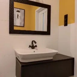 Nowoczesna łazienka z białą umywalką nablatową na czarnej szafce, czarne lustro w ramie z fakturą, geometryczna lampa wisząca i żółty akcent kolorystyczny na ścianie.