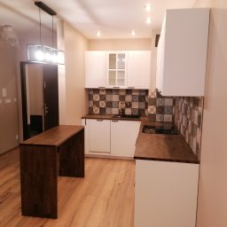 NeoMeb - Nowoczesna kuchnia z białymi szafkami, drewnianym blatem i wyspą w kolorze ciemnego drewna, wzorzysta mozaika nad blatem, drewniana podłoga, oświetlenie sufitowe.