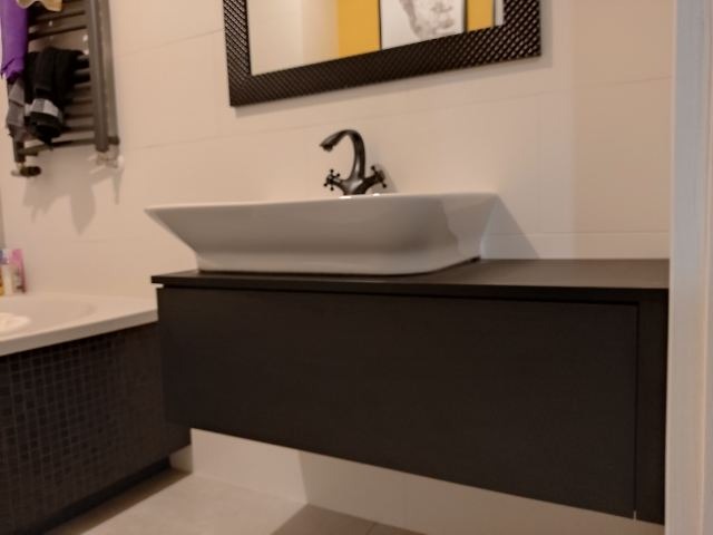 Biała umywalka nablatowa o zaokrąglonym kształcie z czarną armaturą, osadzona na ciemnoszarej szafce łazienkowej z gładkimi frontami, na tle białych płytek i fragmentu wanny z mozaiką.