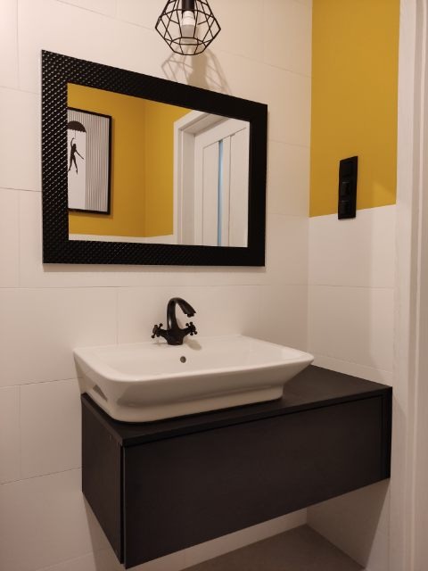 Nowoczesna łazienka z białą umywalką nablatową, czarną szafką pod umywalką, lustrem w czarnej ramie z fakturą oraz żółtą ścianą w tle.