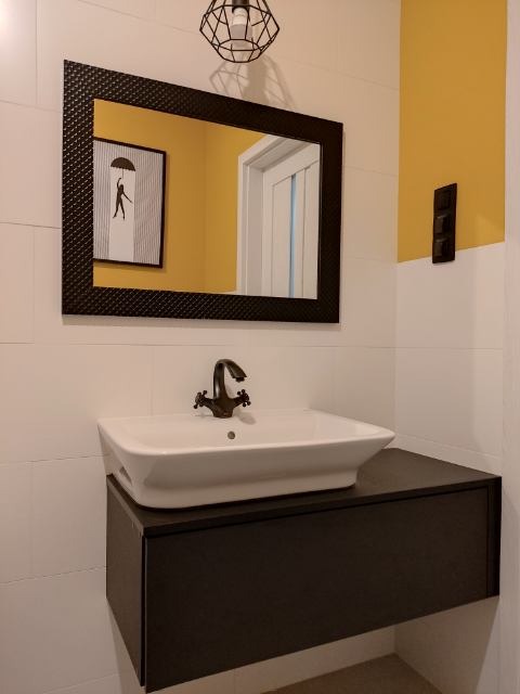 Nowoczesna łazienka z białą umywalką nablatową na czarnej szafce, czarne lustro w ramie z fakturą, geometryczna lampa wisząca i żółty akcent kolorystyczny na ścianie.