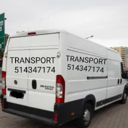 Biały bus Ducato Maxi z napisem 'TRANSPORT' i numerem telefonu 514347174 na boku i tyle pojazdu, zaparkowany na ulicy w pochmurny dzień.