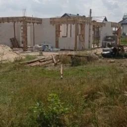 Wznoszenie ścian domu z bloczków betonowych, widoczne drewniane szalunki nad otworami okiennymi i drzwiowymi, na pierwszym planie wysoka trawa i materiały budowlane.